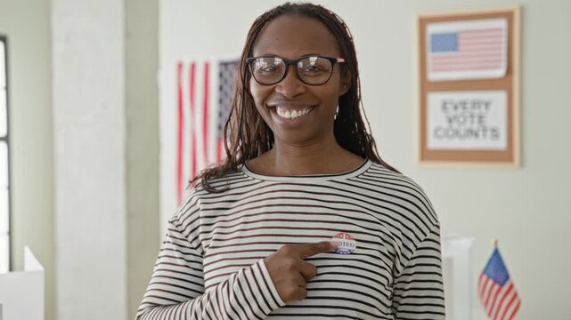Woman points finger to voted sticker on her chest inside a building polling place, smiling with glasses and striped shirt and american flag and poster visible; civic pride participation.