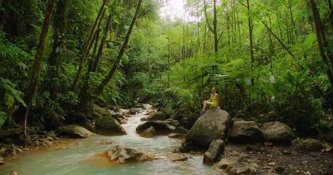 Tourist woman sitting on a large rock by a jungle river in Costa Rica during journey through wold scenic landscapes