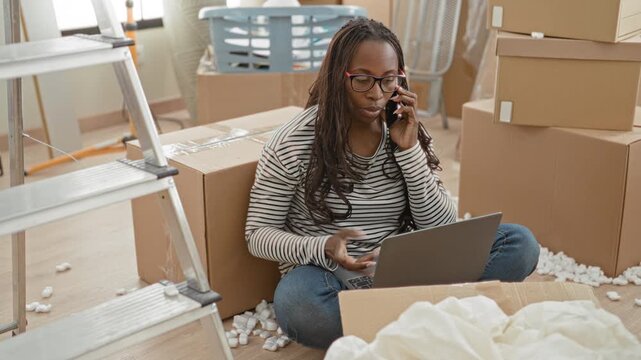 Woman sitting among packed moving boxes holds smartphone to ear and types on laptop in a building with ladder, laundry basket and packing peanuts scattered on floor; stressed relocation.
