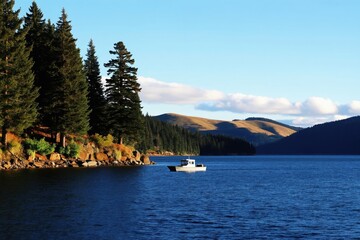 Obraz premium Boat floats on calm blue water near forest shore with rocky bank. Sunlit hills rise in distance under clear sky. Serene natural landscape offers peaceful outdoor recreation.