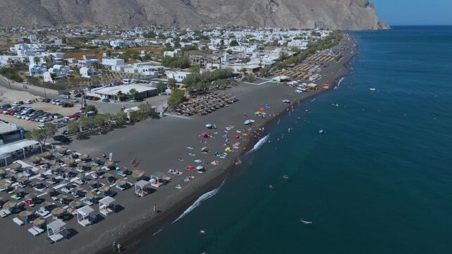Aerial View of Perissa Black Sand Beach Resort and Coastal Promenade in Santorini Greece. A long stretch of dark volcanic black sand lined with organized rows of thatched umbrellas.
