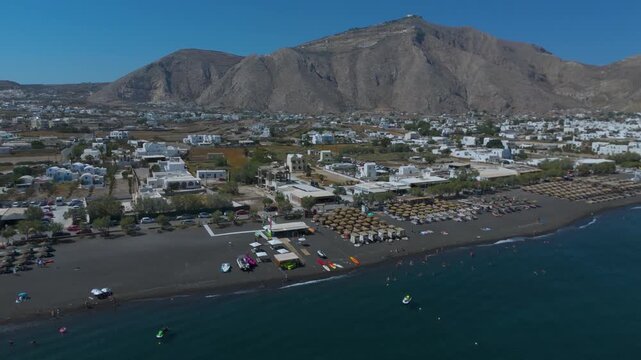 Aerial View of Perissa Black Sand Beach Resort and Coastal Promenade in Santorini Greece. A long stretch of dark volcanic black sand lined with organized rows of thatched umbrellas.