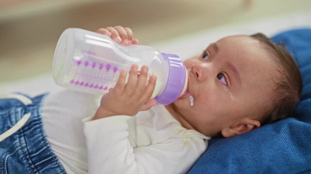 Baby boy drinking milk from a bottle, holding it with both hands as milk drips at mouth while lying on a bed in a bedroom; distress.