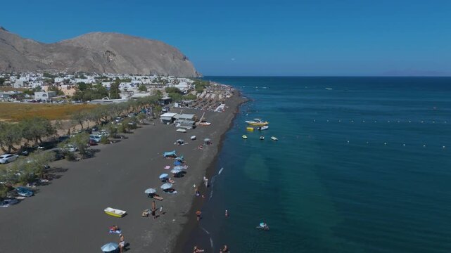 Aerial View of Perissa Black Sand Beach Resort and Coastal Promenade in Santorini Greece. A long stretch of dark volcanic black sand lined with organized rows of thatched umbrellas.
