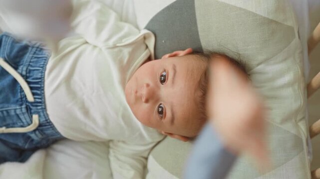 Baby boy lying on bed in building, eyes gazing up at hanging mobile above cradle while wearing white onesie and denim pants; calm curiosity.