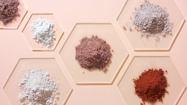 From-above capture of piles of different color powders beige and brown on hexagonal watch glass pieces on a tan-colored background lit with orbiting light casting moving shadows
