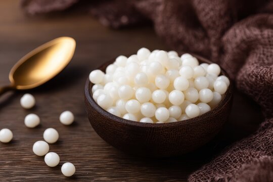 A bowl of white tapioca pearls on a dark wooden table with a golden spoon and burlap fabric