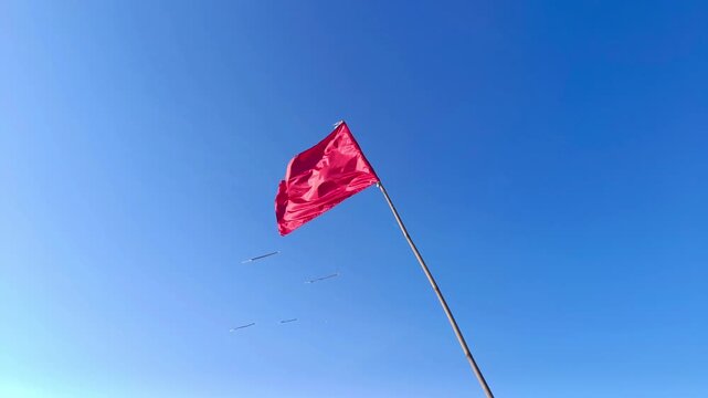 Footage of a rectangular red flag fluttering in the wind on a bamboo pole. The flag is set against a clear, cloudless blue sky, providing a sharp contrast to the red of the flag.