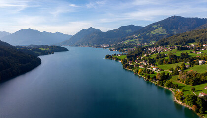Aerial view of a serene mountain lake with lakeside buildings, surrounded by forests and rolling hills, capturing the beauty of a peaceful European landscape. © NafiliaEmil