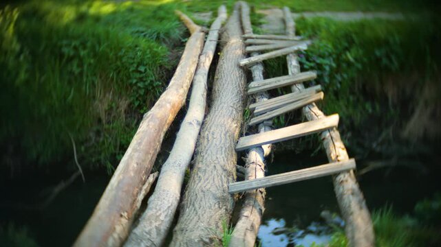 A rickety old wooden bridge spans across a small forest river. Green grass and wild vegetation grow densely on the riverbanks. Cinematic look with vignette and swirled background.