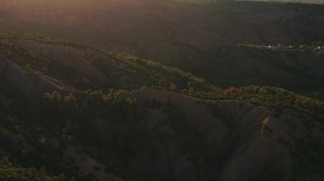 Forested ridge overlooks terraced slopes in Ourika Valley, Al Haouz Province, Marrakesh-Safi, Morocco, illuminated by low sun across High Atlas foothills, drone parallax descent with tilt up shot.
