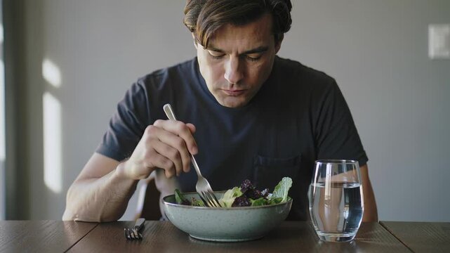 A man eating a bowl of healthy and organic salad in a clear transparent bowl with glass of water while sitting at a table in a white kitchen background
