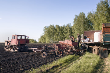 Fototapeta premium Workers Loading Seeds and Nitrogen Fertilizer into Seed Drill Near Tracked Tractor During Field Preparation for Sowing Operations
