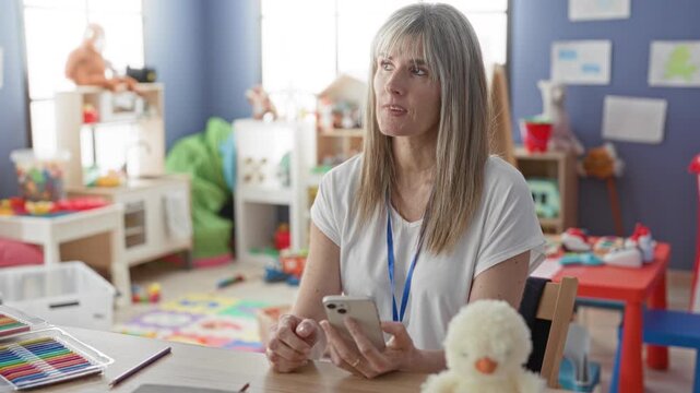Blonde woman teacher holds smartphone, fingers tapping screen and glancing up while seated at a table with art supplies and a stuffed chick in a preschool classroom building; thoughtful reflection.
