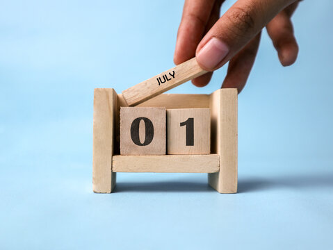 A woman's hand holds a wooden block with "July" written on it on a wooden block calendar for July first against a light blue background.	