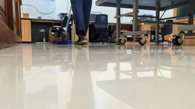 A janitor sweeps the office floor, keeping the workspace clean and tidy as the polished surface reflects the room and surrounding furniture.
