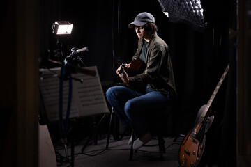 Fototapeta premium A woman plays an acoustic guitar in a dark studio, surrounded by music stands, microphones, and other guitars in the background.