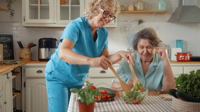 Smiling caregiver and senior woman laugh and chat while preparing a healthy salad together in a cozy home kitchen.