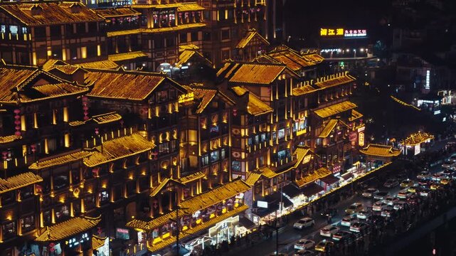 Elevated wide shot of heavy traffic moving past illuminated traditional buildings at Hongyadong Folk Custom Scene Area in Chongqing at night, with glowing rooftops and dense urban skyline