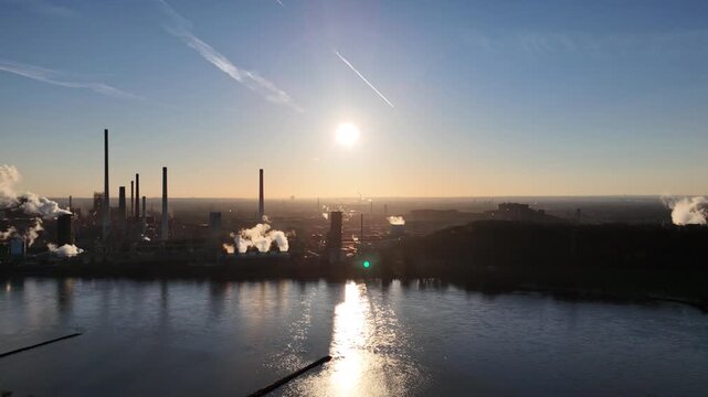 Backlit drone move along the opposite riverbank with sunlight and industrial shadows shimmering on water