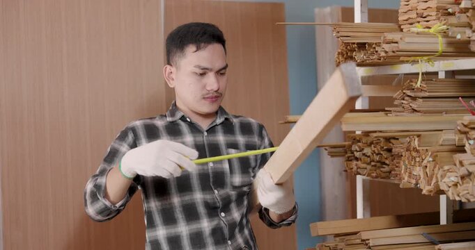 Asian adult male woodworking business owner pulling tape measure across vertical wood board, verifying size proportions during material selection process inside interior furniture material store
