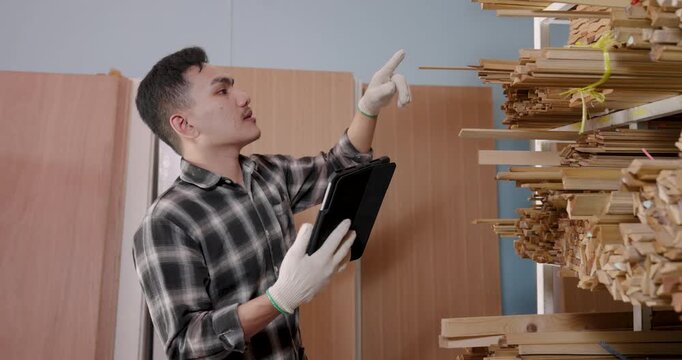 Asian male carpenter pointing upward toward stacked wood strips, visually estimating length and thickness while holding tablet, planning material selection for furniture manufacturing in workshop