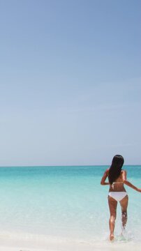 Vertical video of excited young woman in white bikini running into water on beach. Cheerful female is enjoying nature during her summer vacation travel.