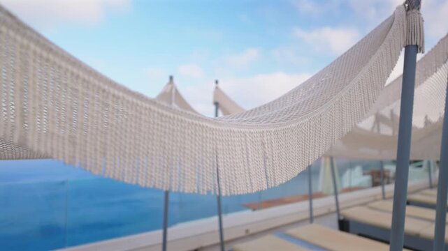 Elegant white woven hammock with decorative fringe sways gently at a luxury resort terrace in Santorini Greece. Sun loungers and parasols create a serene poolside atmosphere with blue sky.
