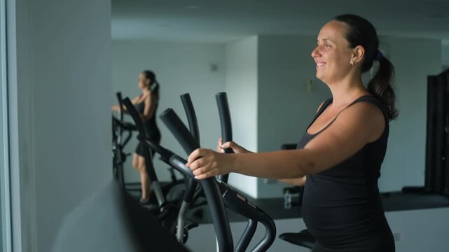 Young woman in black sportswear exercising on an elliptical trainer machine in a bright modern gym interior with large windows. Fitness concept