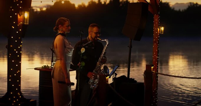 Female wedding singer and saxophonist performing on lakeside wooden stage at dusk