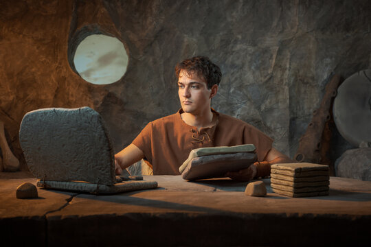 Funny Serious Young Prehistoric Caveman Working with Stone Laptop and Tablet Document in a Cave