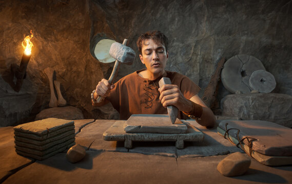 Funny Young Prehistoric Caveman Carving Stone Tablet with Chisel in a Cave