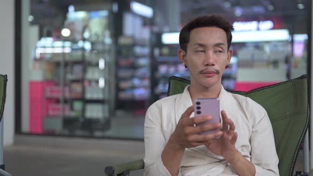 Thai Non-Binary Gay Man Sitting Outdoors and Texting on Smartphone at Night