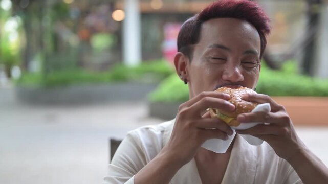 Thai Non-Binary Gay Man Eating and Biting a Hamburger Outdoors Horizontal Shot