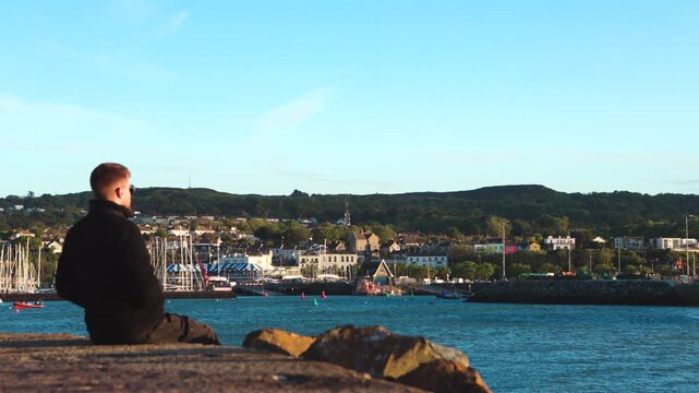 Man Sitting at Howth Harbour Watching Boats and Village During Windy Sunny Evening