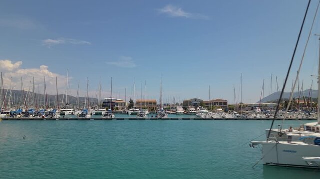 Drone footage simulating a yacht leaving its berth and navigating through a marina channel between docked sailboats and catamarans in Lefkada, Greece.