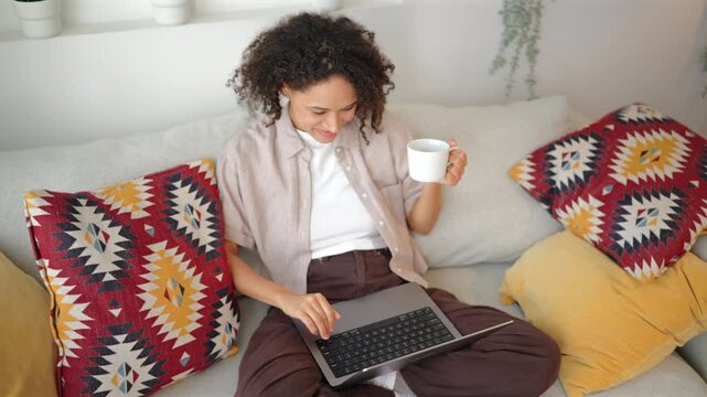 African American young women with coffee working on laptop during remote working job. Diverse girl sits on cozy sofa with colorful pillows browsing computer screen and enjoying hot beverage at home.