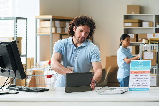 Young man processing orders on tablet at pickup counter while Asian woman sorting parcels in delivery hub. Ideal for logistics service, ecommerce fulfillment marketing
