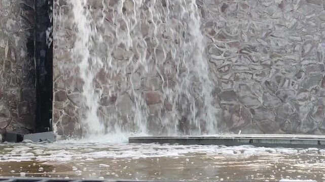 Stone wall channels flowing water into a pool below. Water cascades down rugged, textured surface. Scene suggests tranquil, natural design in urban setting