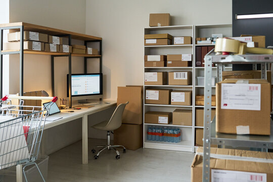 Wide shot of order pickup workspace displaying packed boxes, shipping labels, computer station, shopping cart in fulfillment center. Supporting e commerce logistics, delivery service marketing