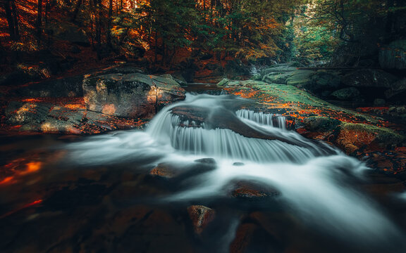 View of a small waterfall cascading over dark rocks surrounded by autumn forest with orange leaves in Szklarska Poreba, Lower Silesian Voivodeship, Poland.