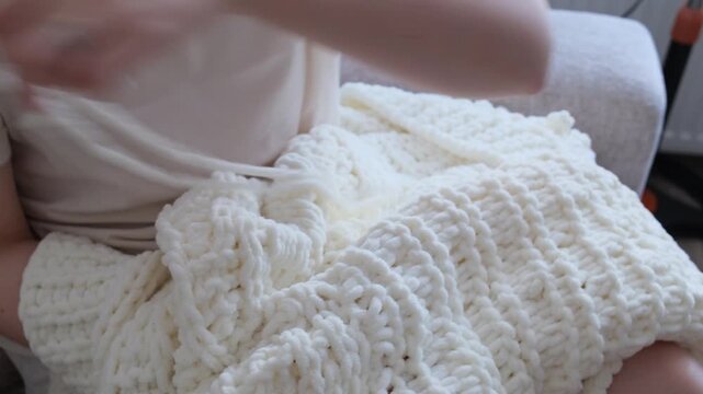 Close up of female hands knitting a cozy, soft white cardigan using chunky plush loop yarn at home.