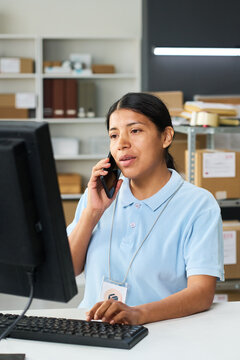 Vertical shot of young Latina woman managing order pickup desk, speaking on smartphone and using desktop computer. Supporting delivery logistics, retail service, ecommerce operations