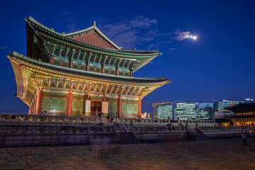 Fototapeta premium Stunning night view of Geunjeongjeon Hall at Gyeongbokgung Palace with full moon and modern city skyscrapers background in Seoul South Korea
