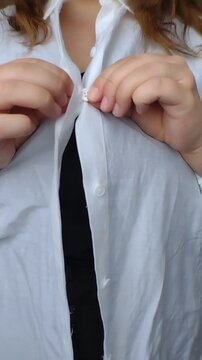Close-up of a plus-size woman's hands buttoning up a white cotton shirt over a black top. Vertical video for body-positive casual fashion retail, daily routine, and commercial apparel ads.