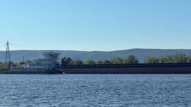 Cargo Ship on the River: A large cargo ship with a long row of barges navigates a wide, calm river under a clear blue sky, with rolling hills and scattered trees in the background.