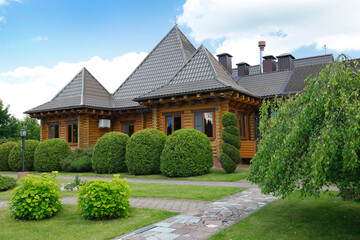 Obraz premium Beautiful wooden log house villa with perfectly manicured green topiary shrubs on sunny day. Luxury rustic timber cottage architecture with decorative spiral evergreen bush and neat stone pathway