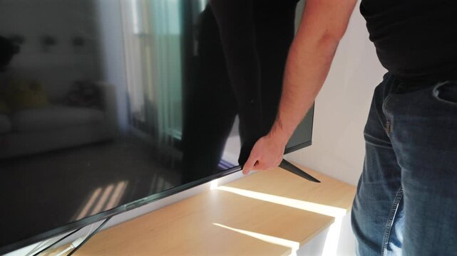 Movers carrying television at home as man carefully positions large flat screen TV on wooden media console. Bright living room with natural sunlight streaming through windows during furniture.