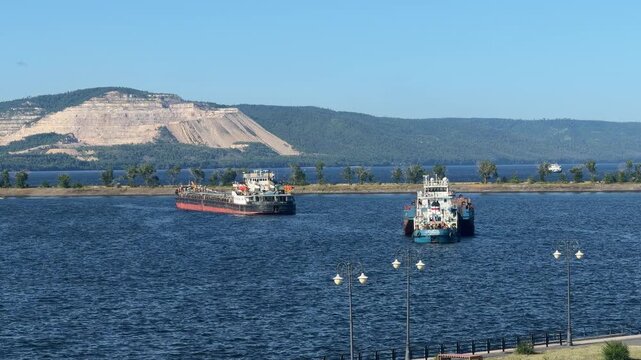 Barge Traffic on Wide River: Two barges navigate a vast, blue river on a clear day, with a forested shoreline and a distant, mountainous landscape under a bright sky.