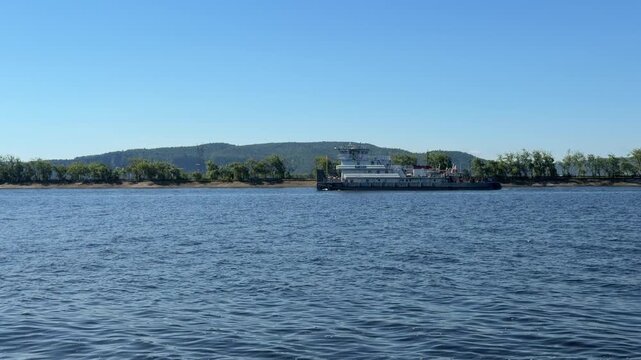 Ferry Boat on the River Volga: A large ferry boat navigates across a wide, calm river under a clear blue sky, with a tree-lined shore and rolling hills in the background.
